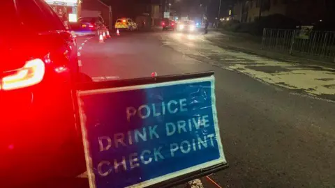 Cambridgeshire Police A road at night, featuring a blue police sign, orange cones and vehicles parked at the roadside