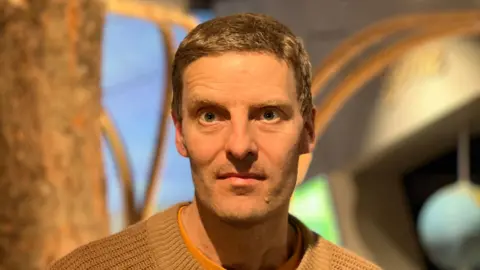 Artist Charlie Winney. He is clean shaven with sort brown hair and is wearing a light brown jumper