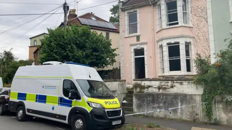 A white police van with blue and yellow hi-vis decals, parked beside the pavement on a residential street. There is blue and white cordon tape set up next to it, securing off a faded pink house with bay windows and a dilapidated garden wall.