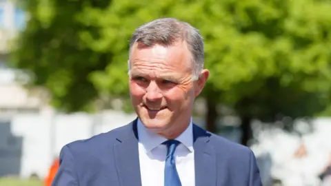 Richard Evan Rhys Williams pictured leaving Cardiff Crown Court. He smiles through the sunshine, squinting slightly. He has grey short hair and wears a navy suit, white shirt and navy tie. 