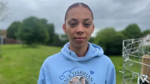 A young woman stands in a park with green grass and trees behind. She's wearing a blue hooded top with the name and pictures of her brother on the front, with blue hearts. To the right of the screen there is a fence with fairy lights on. 