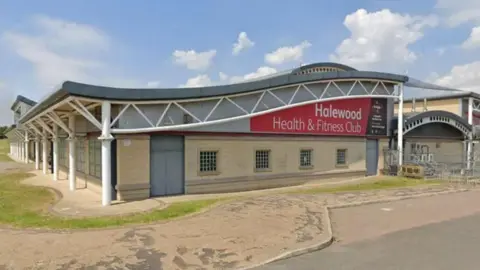The outside of Halewood Health and Fitness Centre is a large building with a roof in the shape of a wave in the sea.