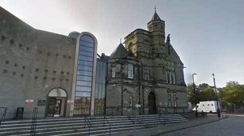 A general view of Kirkcaldy Sheriff Court. An older building made of dark stone is connected to a newer building, made of lighter stone, by several glass panels. A large brick archway is in the middle, which is also filled with glass panels.