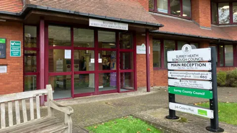 BBC/Jack Fiehn Entrance to Surrey Heath Borough Council in Camberley. It is a red-brick building with a wooden bench outside and a path and a grass verge.