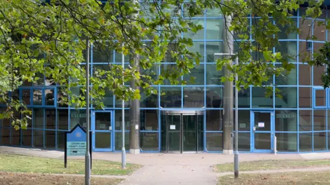  John Fairhall/BBC Basildon Crown Court can be seen through green trees. It has many square panelled windows with blue frames.