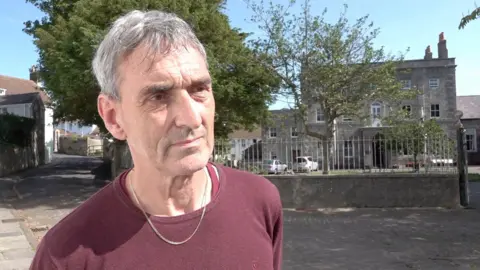 Photo of a man with short grey hair wearing a burgundy long-sleeve top with a thin gold necklace.