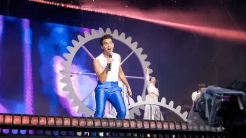 Paul Campbell Mika, dressed in a white vest and electric blue trousers, on stage at Bella. His set design included large clock cogs.