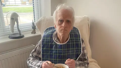 Terry sits in an armchair eating a bowl of cereal. He has an apron around his neck and white hair.