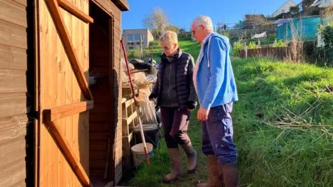 Paul and Angela Rodgers are walking into their wooden shed on the allotment. They are both wearing wellies and are walking along some grass towards the open shed door. There is a red hoe resting on the wall of the shed. The sky is blue and it is sunny.