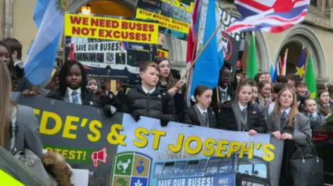 Pupils in school uniform hold up banners and flags. One banner reads "we need buses, not excuses" while another says "stop brutal school journey cuts".