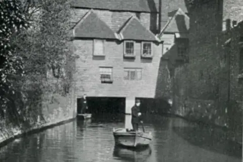 Eastbridge Hospital A medieval building bridging over a river, in black and white. Two punters are seen on the water, with trees on the wall banking the water.