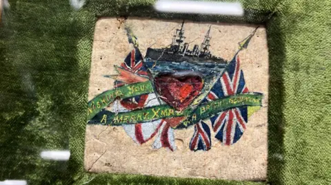 Leeds Museums The biscuit features a colourful hand-painted design. It shows a red heart flanked by two Union flags on either side. Behind the heart is a naval warship. There is a message on the surface which partly reads 'Wishing You A Merry Xmas'.