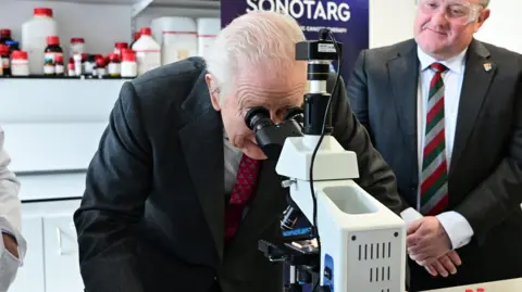 PA Media King Charles who has grey hair swept bac from his head and is wearing a dark suit and red tie looks through a microscope at Ulster University. A man also wearing a dark suit and wearing safety goggles stands beside him