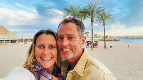 The image shows a couple posing for a selfie; palm trees and a beach can be seen behind them.