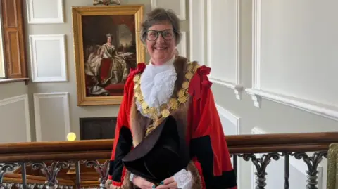 LDRS A lady wearing mayoral garb including a red cape and gold chains stood in a council office. She has short grey hair and is smiling and wearing glasses.