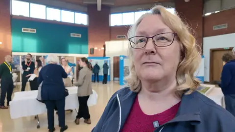 A woman with mid length wavy hair and glasses in a blue jacket stands in the hall containing the reservoir exhibition