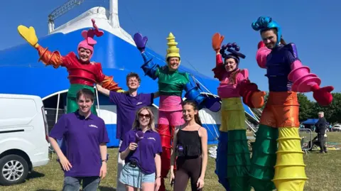 Four people on stilts in rainbow coloured circus costumes stand and hold their arms up, while three BBC reporters wearing purple T-shirts stand in front of them. They are in a field with a large blue marquee behind them.