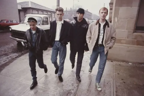 Getty Images The four members of Wet Wet Wet walk along the street in Glasgow, four abreast in 1987. It's a typical Glasgow street with tenement flats in the background, cars parked on wither side of the street. The four wear casual clothes and all wear jeans. It is a drizzly day.