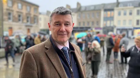 A dark haired man with a brown jacket, pink shirt and blue tie looks at the camera.