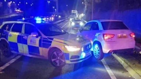 It is night time and a blue lit police car is parked in behind a white car in order to get it to stop. There is another car with its headlights on driving towards the two stationary cars.
