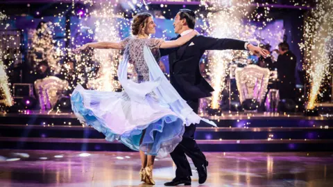 BBC/PA Karen Carney and Carlos Gu dancing with their arms wide, smiling at each other. Carney wears a clitter light blue dress while Gu is wearing a black suit with his black hair slicked back.