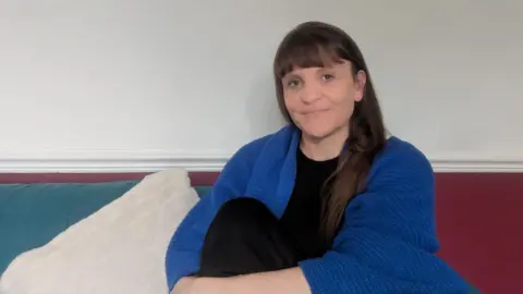 Tonia Dunn Person with long straight hair and bangs sitting on a sofa against a light-colored wall, wearing a blue cardigan over a black top, with a white cushion to one side