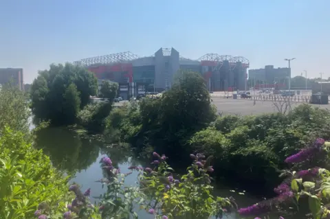 Photograph of Old Trafford, with the Bridgewater Canal in the foreground. The image also show some of the stadium car parks.