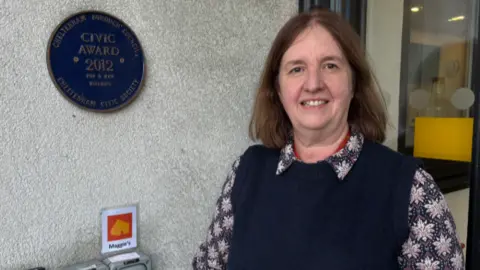 A photo showing Nicky Peregrine, centre manager at Maggie's charity in Cheltenham. She is stood next to a wall which has a blue plaque on it. She is wearing a blue sleeveless jumper and a patterned black and white shirt