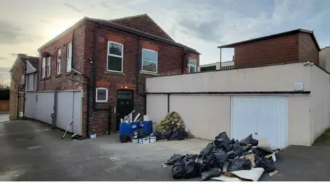 LDRS A two-storey red brick building with a cream-coloured single-storey garage/store room attached. Two piles of building waste materials lie in the yard next to the buildings.