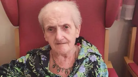 Dudley Metropolitan Borough Council A woman with short grey hair and wearing a blouse, sits on a large red chair. She is also wearing a necklace.