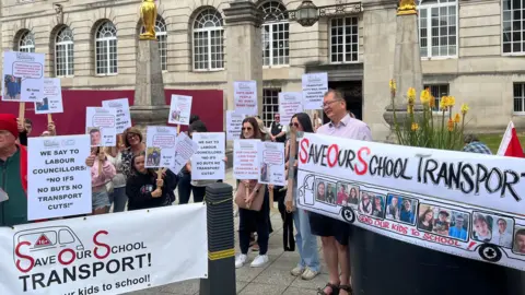 A group of parents with banners and posters in front of Leeds Civic Hall. Banners say 'Save Our School Transport' and have images of children.