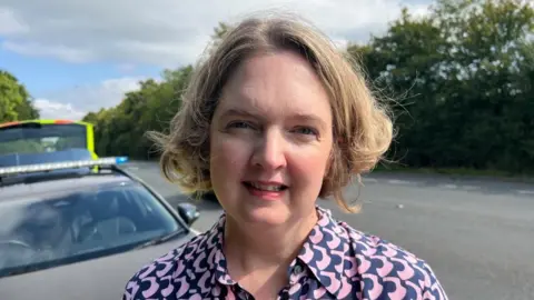 A woman with mid-short blonde hair standing by a police car on a road.