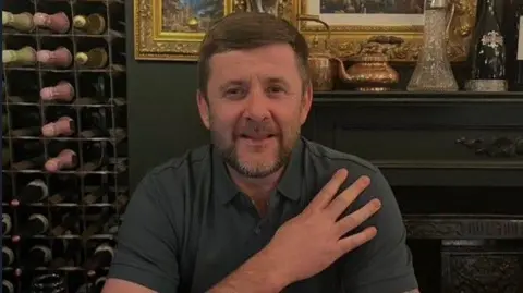 A man seated at a dining table in what appears to be an upscale or traditional-style restaurant or dining room. He is wearing a short-sleeved, dark-coloured polo shirt, and has brown hair with a beard.