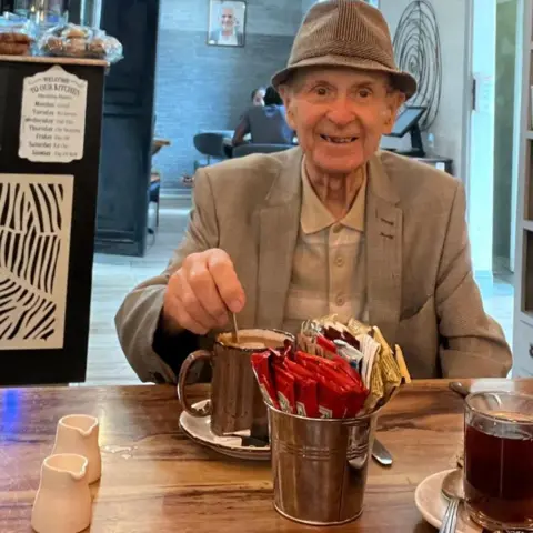 Mackey family An elderly man smiles (right) for a photo in a cafe. He is wearing a pork-pie hat and a cream jacket and light shirt. He is stirring a hot drink in a mug on a table in front of him which contains a mental container of condiments
