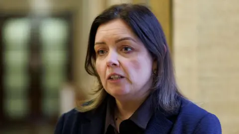 PA Media Deirdre Hargey is standing in Stormont's Great Hall. She has dark hair and is wearing a navy jacket. 