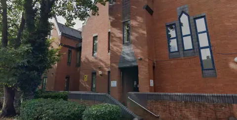 Google A red brick building with trees and a bushes outside and reflective windows.