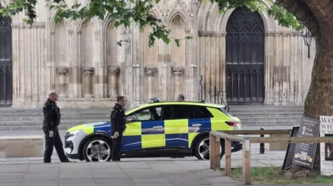 Actors dressed as police officers stand around a police car parked outside York Minster.