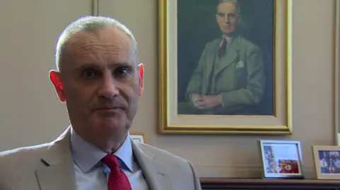 Nick Coburn sitting in an office with an official portrait of someone behind him. There are some smaller, family-style pictures in frames on the right of the picture, atop a table. Mr Coburn is wearing a beige suit and red tie. He has cropped grey hair.