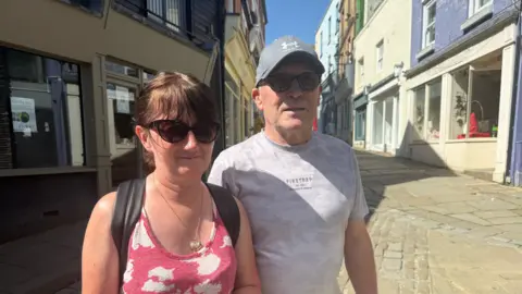 Donna Daly and James Marrin are pictured standing outside in a shopping area with a narrow street behind them. Donna is wearing a pink vest top, and James is wearing a T-shirt. Both have sunglasses on and look hot. There is a clear, blue sky.