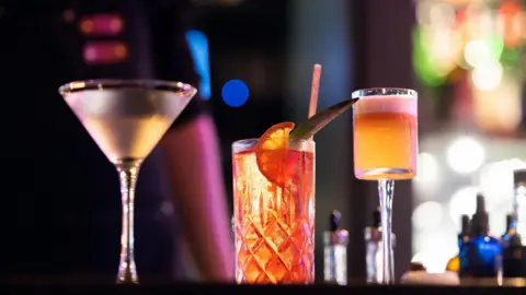 Getty Images Three cocktails sit on a bar counter in a darkly lit bar. The cocktails are illuminated, a martini glass slightly out of focus on the left, and two other drinks in focus to the right. In the background is the bartender's arm, and the bright lights of the bar shelves.