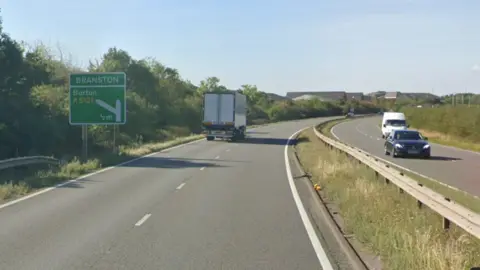 A two lane carriageway on the A38. Traffic can also be seen heading in the opposite direction. There is a green A road sign on the left that indicates an upcoming slip road towards Branston. A van is travelling towards the junction