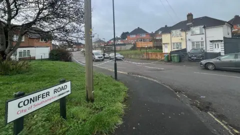 A shot of Conifer Road. Police tape can be seen blocking off access to the street.