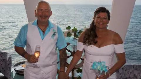 Villa Vie Residences A couple on a cruise ship smiling. The man (left) wears a blue shirt and white waistcoat. The woman (right) wears a white dress and carries blue flowers. She has curly brown hair. The sea is visible behind them.