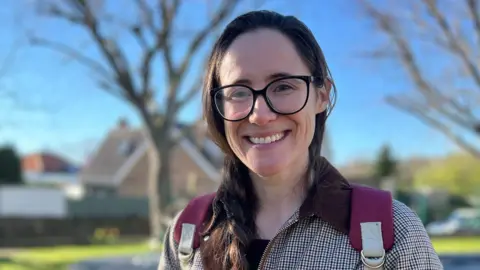 Sinead has braided hair and stands on a grassy area with bare trees and houses behind them. She is wearing a checked coat and a backpack under a bright blue sky.