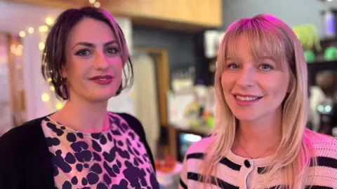 Madelaine Thomas and Jess Davies in a coffee shop smiling at the camera. Madelaine has dark short hair and is wearing an animal print top and black cardigan. Jess has long blonde hair with a fringe and us wearing a black and white striped cardigan. It is a head and shoulders shot of the two of them.