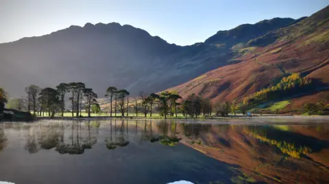 A mountain ridge sits in front of a blue sky and drops steeply down to a lake which is fringed by a number of trees. The mountainside and trees are reflected in the dark water