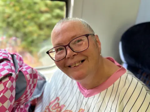 A photograph of passenger Sharon Buttler who is seated on the train and is wearing a white top with pink trimmings and blue stripes. She has grey hair, which is pulled back, and glasses.