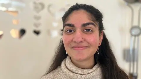 A close-up of Mehreen Ahmed who received a heart transplant in June 2023. She has long dark hair, wears a cream polo-neck sweater and is smiling into the camera. There are heart decorations behind her.