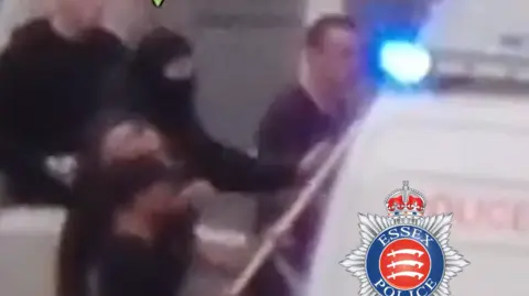 Essex Police A blurry still from a video showing Fleming, still wearing his black coat and face covering, hitting the side of a white police van with its blue lights on.