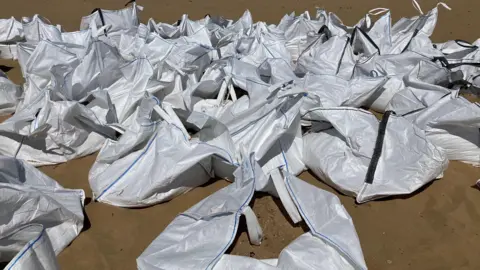A close up of a stack of large white bags filled with sand containing tiny white pellets, lying on a beach.
